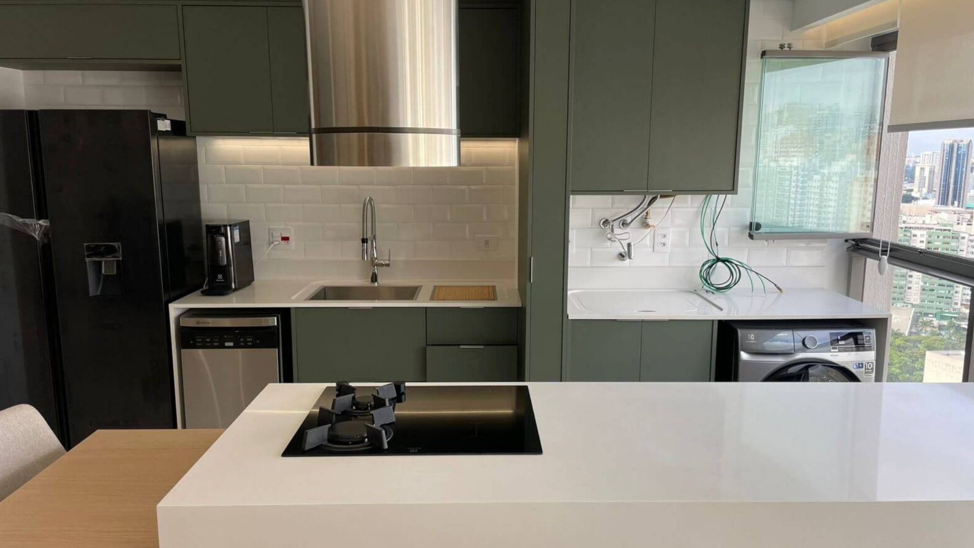 Cozinha reformada de casa de alto padrão em São Paulo, armários planejados em tom verde oliva, bancada branca com cooktop integrado, coifa em inox, revestimento em subway tile branco, vista para a cidade, fotografia realista com luz natural