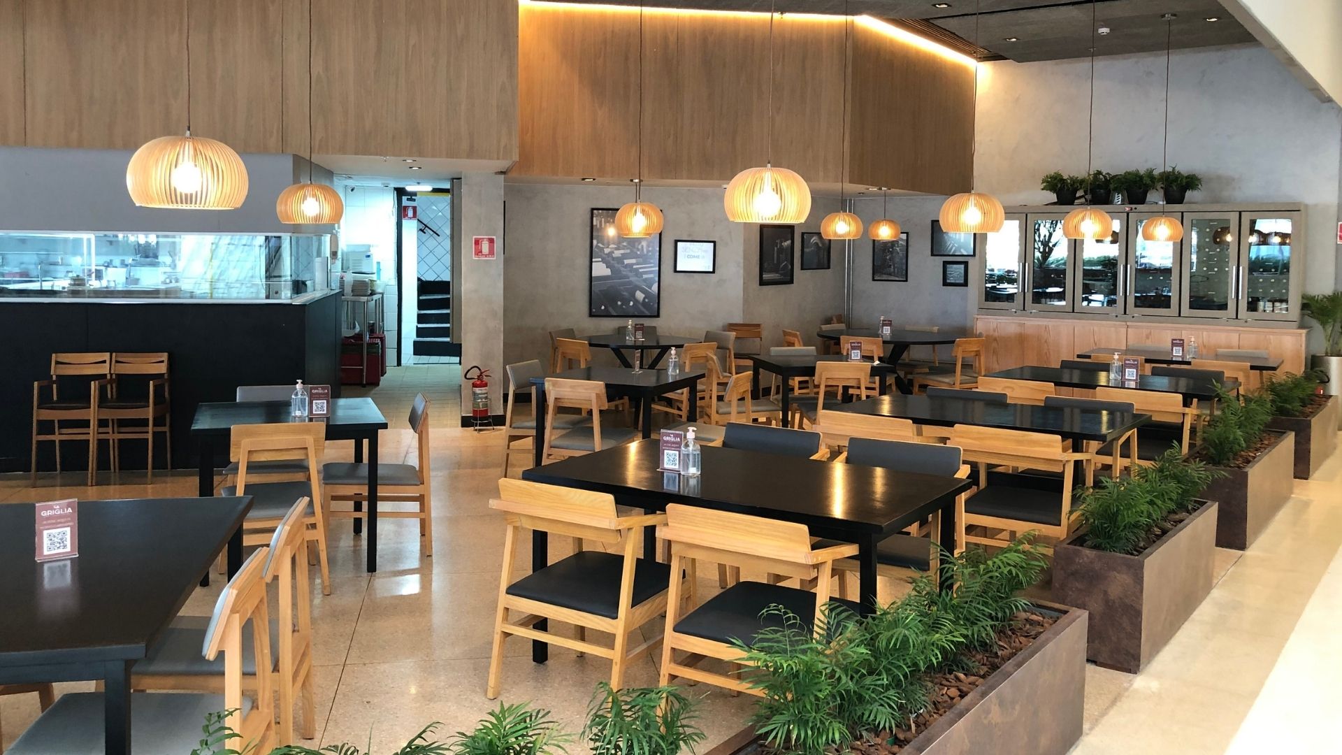 Salão de restaurante comercial reformado em São Paulo, mesas em madeira e cadeiras estofadas, pendentes decorativos em ripas de bambu com iluminação quente, paredes em revestimento cimentício, jardineiras com plantas verdes entre as mesas, ambiente aconchegante e contemporâneo, fotografia realista