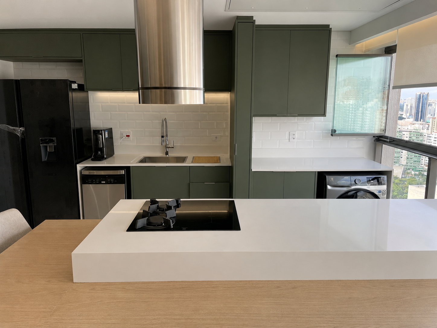 Cozinha planejada de reforma residencial de alto padrão, armários em verde militar, bancada em porcelanato branco, coifa cilíndrica em inox, revestimento em subway tile branco, vista panorâmica da cidade de São Paulo ao fundo, iluminação natural diurna, fotografia realista